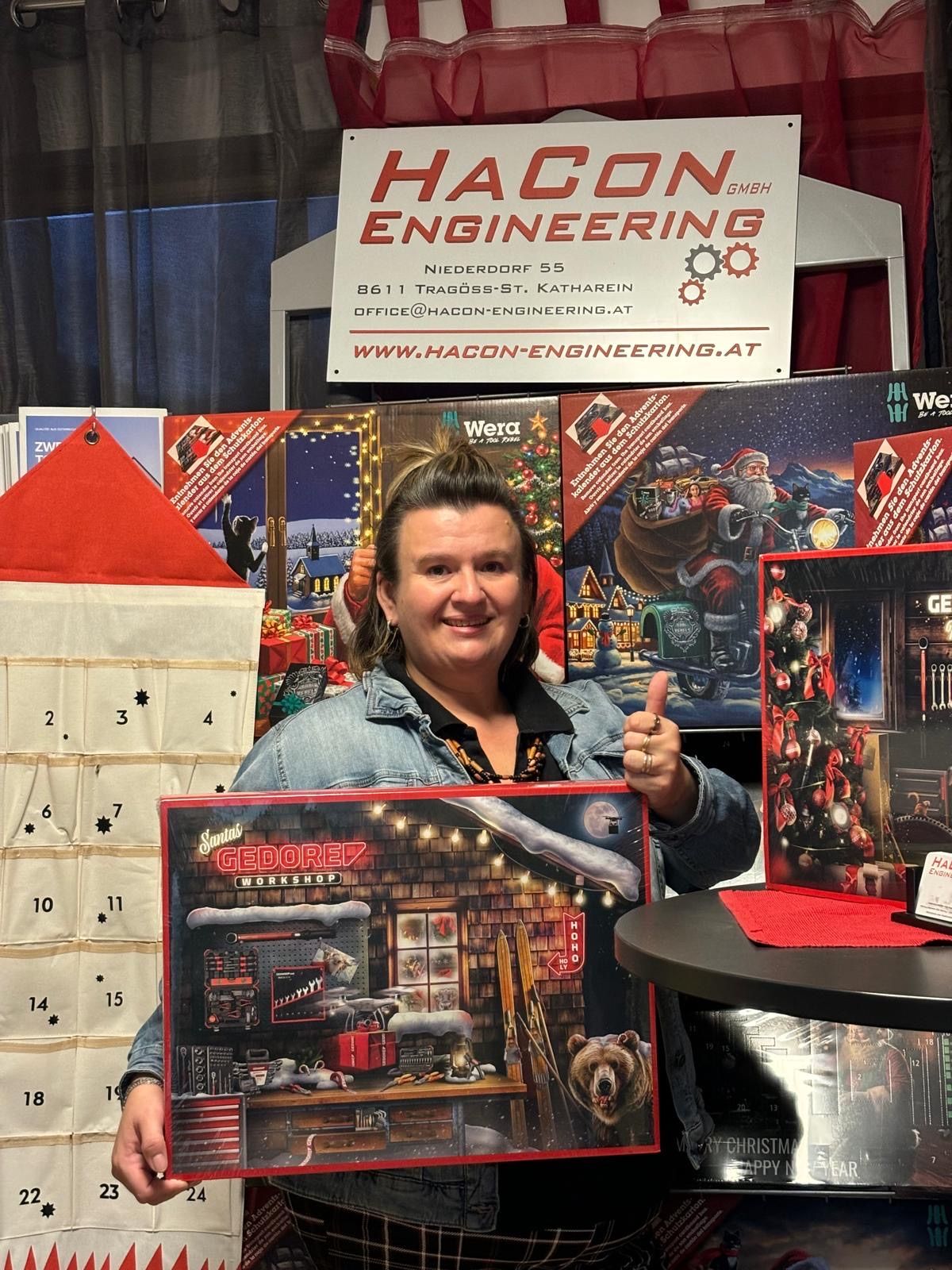 Eine Frau hält einen Gedore-Workshop-Adventskalender vor einem Hacon Engineering-Banner und Weihnachtsdekorationen von Santa Claus.