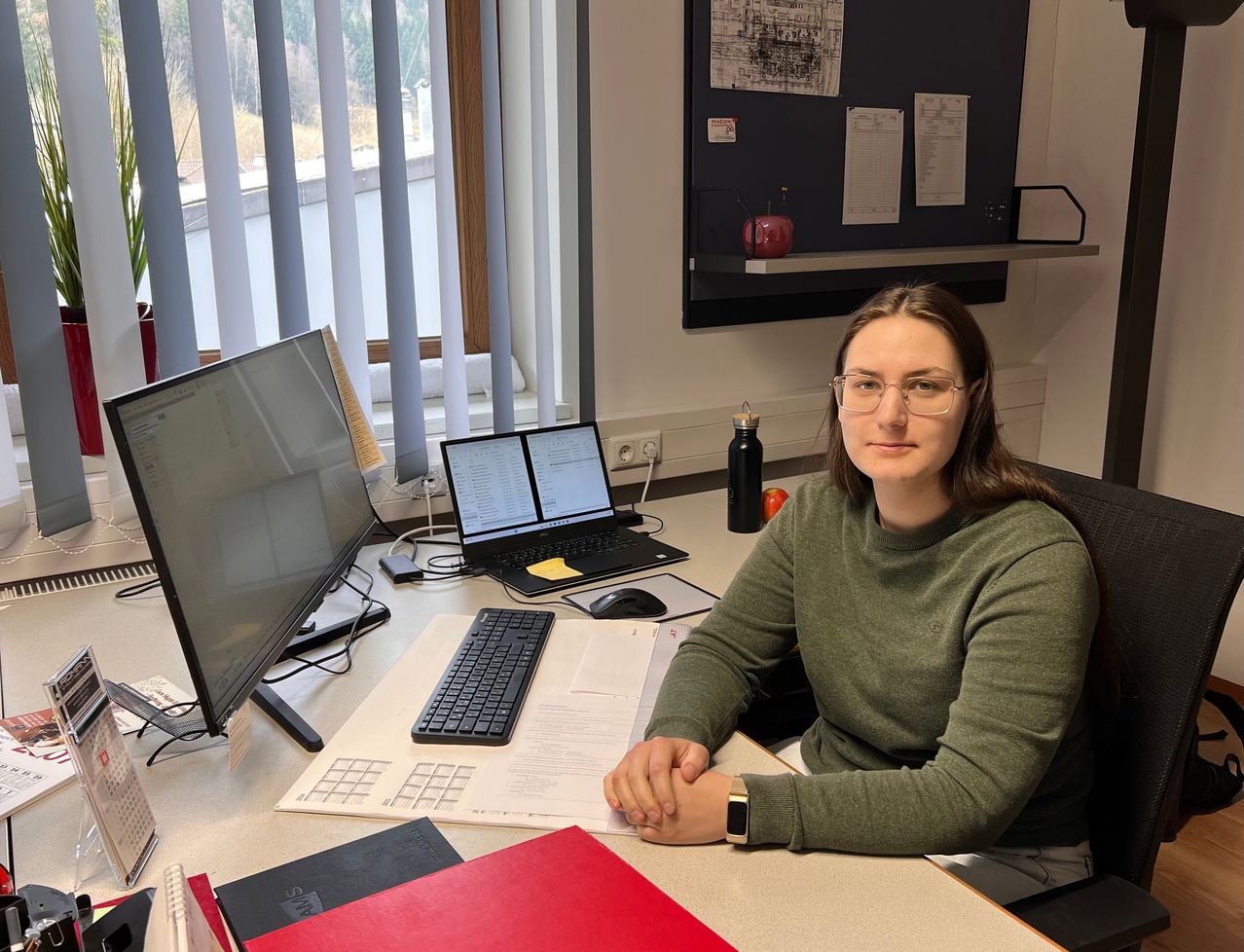 Eine Frau sitzt an einem Schreibtisch mit mehreren Computerbildschirmen, einem Laptop und Papieren. Sie trägt eine Brille und ein grünes Shirt. Hinter ihr befindet sich eine Pinnwand mit angehefteten Papieren.