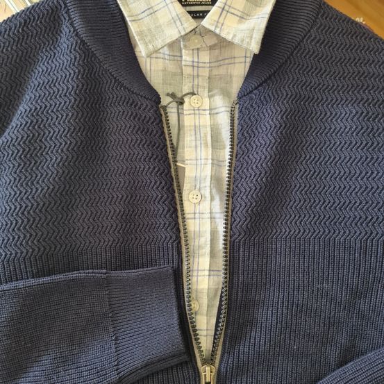 Ein dunkelblauer Schalkragenpullover mit Zickzackmuster und einem teilweise geöffneten Hemd mit Knöpfen.
