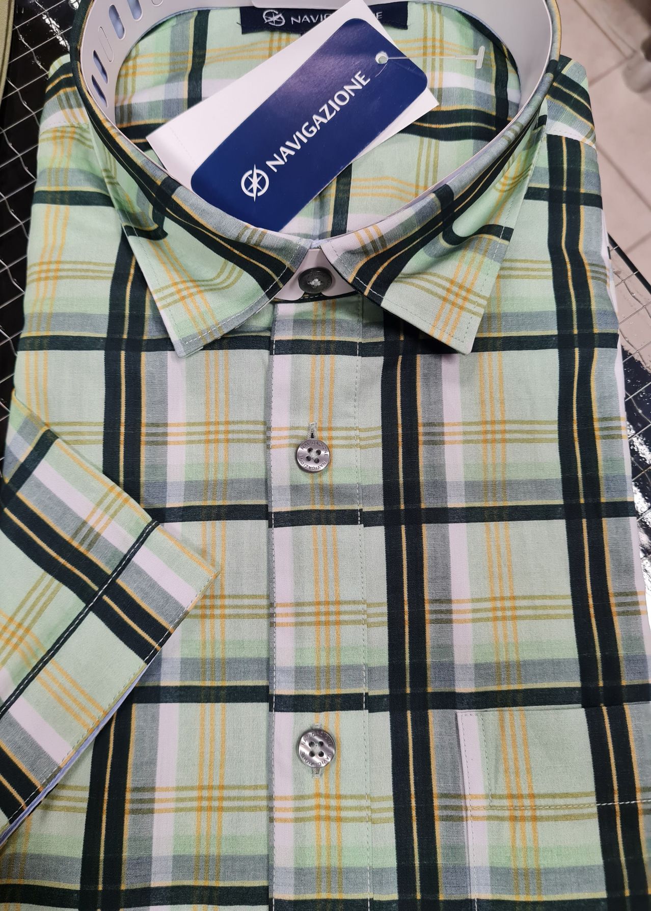 Ein hellgrünes und schwarzes kariertes Hemd mit einem weißen Kragen, Knöpfen und einem blauen Etikett mit dem Wort NAVIG. Das Hemd ist ordentlich gefaltet.