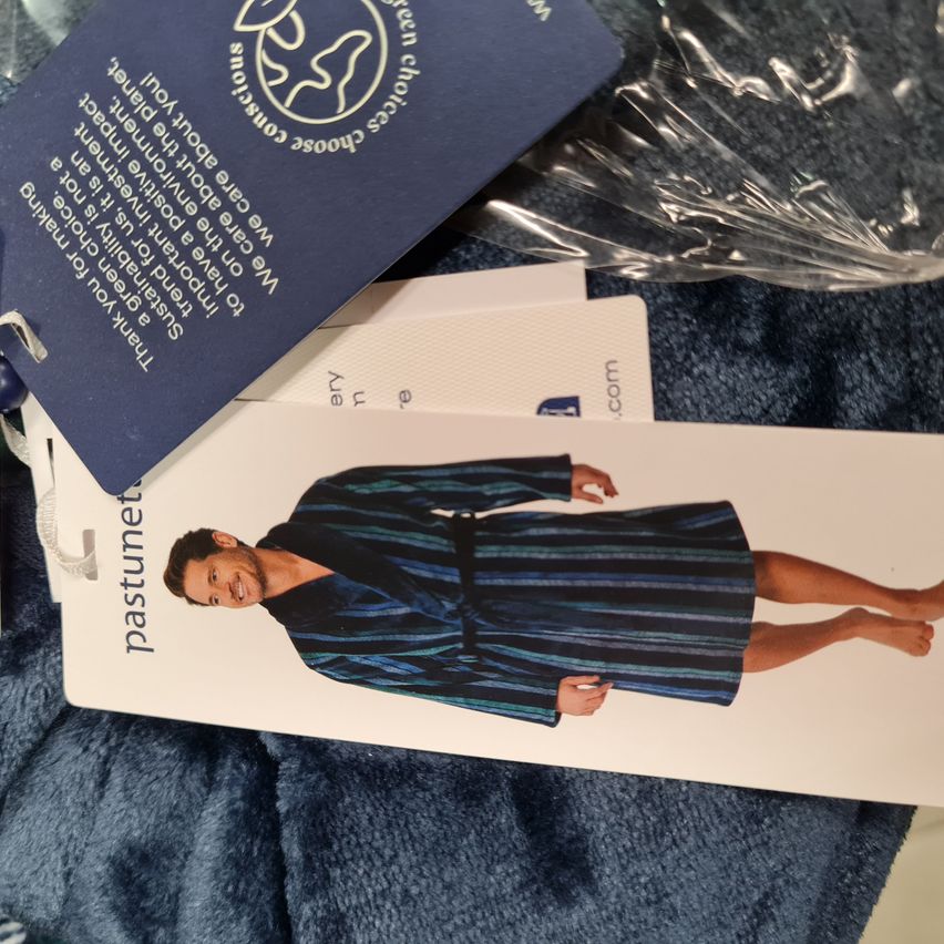 Ein blauer Kleidungsanhänger mit weißem Text auf einer weißen Karte mit einem Bild eines blauen Bademantels. Der Anhänger sagt 'pastunet'. Ein Plastikbeutel ist obenauf.