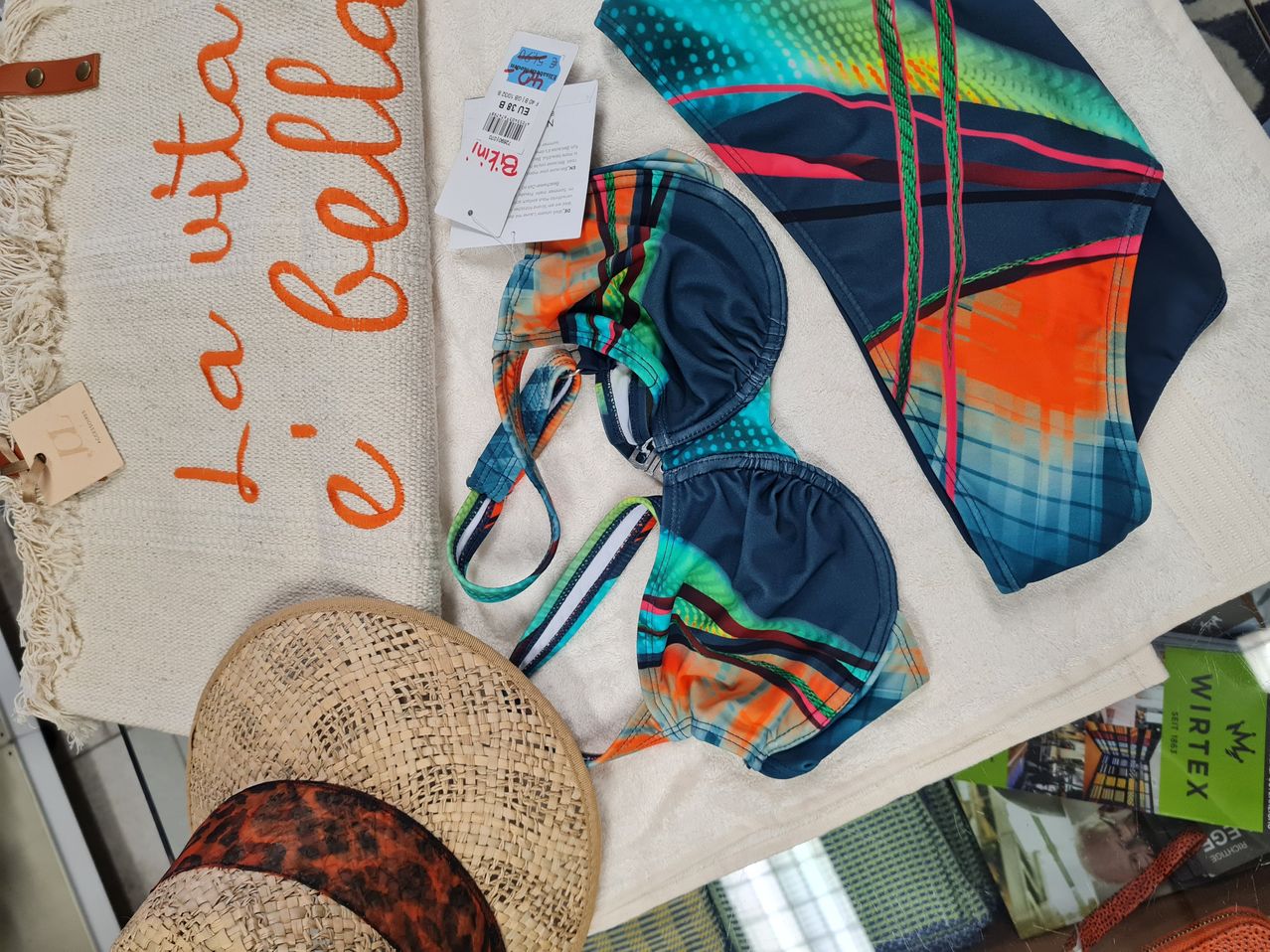 Ein bunter Bikini mit mehrfarbigen Streifen, auf einem weißen Tuch mit einem Etikett, einem Hut und einer Tasche mit orangefarbenem Text.