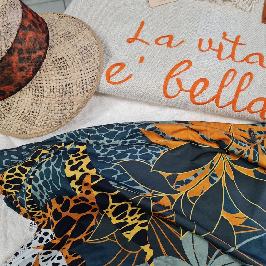 Ein Leoparden-Print-Stoff und ein Strohhut liegen auf einem Handtuch mit dem Text 'La vita e bella' in Orange.