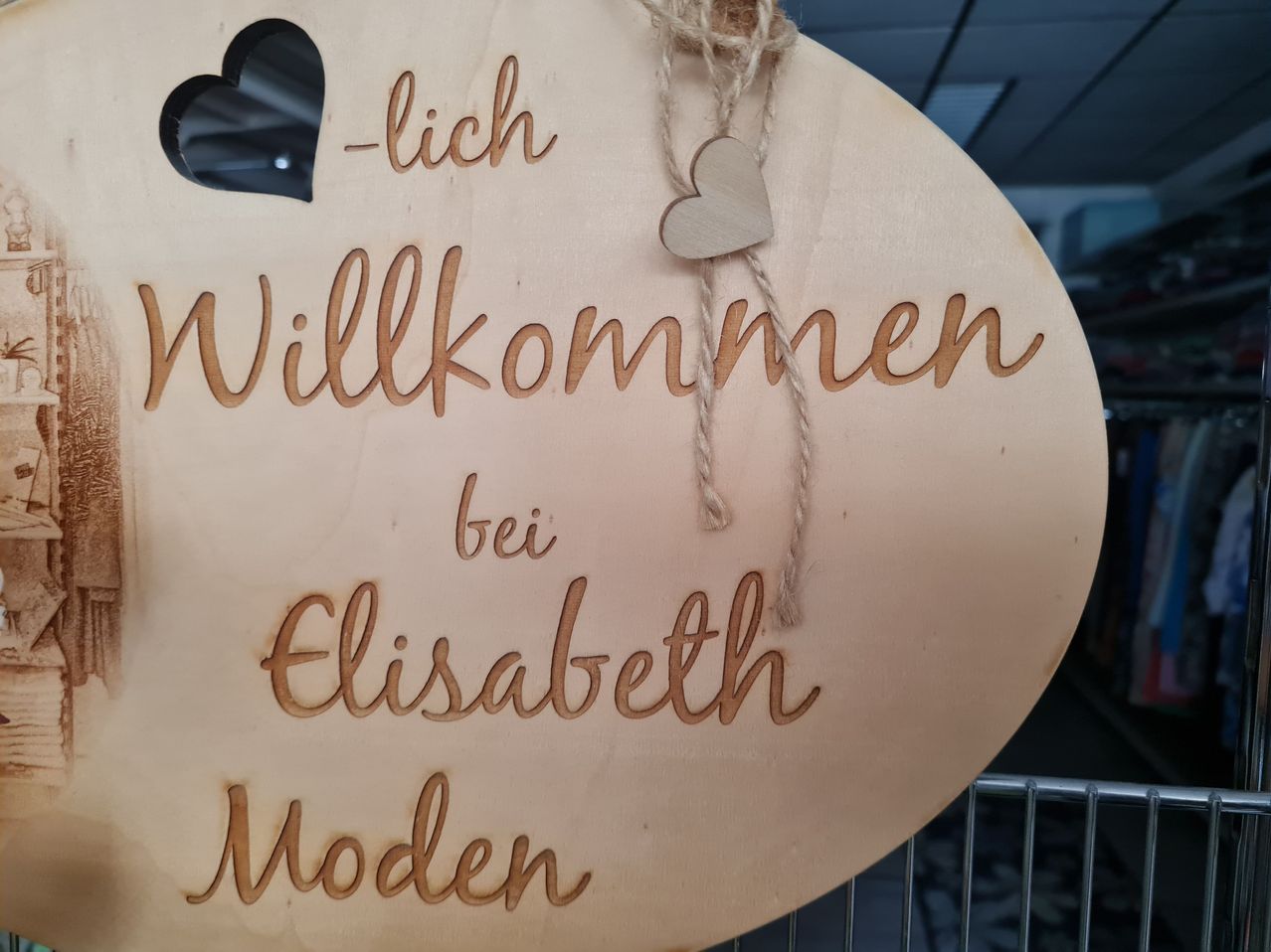 Ein Holzschild mit Herzausschnitt zeigt 'Willkommen bei Elisabeth Moden' mit einem kleinen Herz, das an einem Seil hängt. Das Schild ist an einem Metallregal befestigt.
