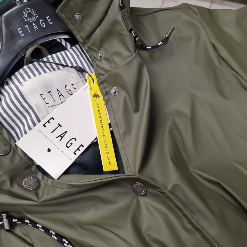 A green jacket with Estage branding hangs on a hanger. Inside, a folded shirt and a yellow tag are visible. The jacket has metal buttons and zippers.
