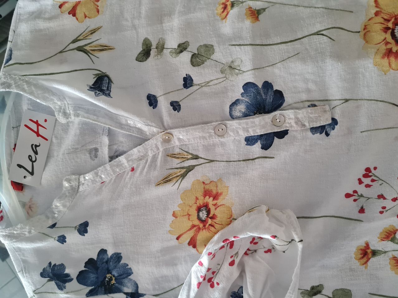 Ein weißer Stoff mit Blumendruck, mit blauen und gelben Blumen, Blättern und Stielen. Der Stoff hat eine leicht strukturierte Oberfläche und scheint gefaltet zu sein.