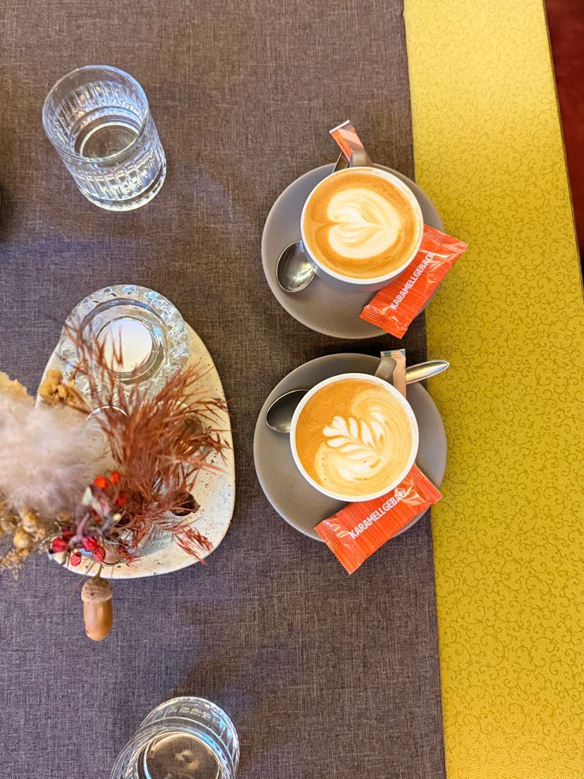 Zwei Tassen Kaffee mit dekorativer Latte-Kunst stehen auf Untertassen mit Löffeln, neben einer dekorativen Schale mit getrockneten Pflanzen, einer Glaskerze und einem Glas Wasser.