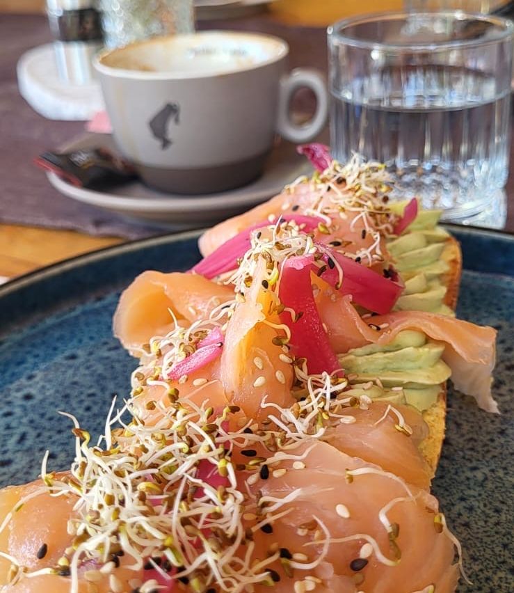 Eine Nahaufnahme eines Tellers mit Räucherlachs, Avocado und Sprossen. Im Hintergrund stehen eine Tasse Kaffee und ein Glas Wasser.