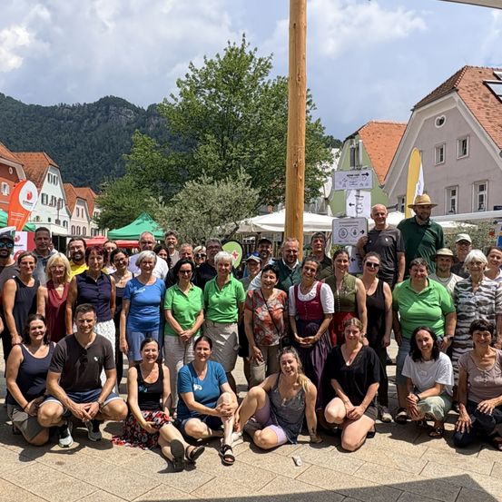 Eine Gruppe von Menschen, darunter Männer und Frauen, posiert für ein Foto in einer Außenumgebung. Sie stehen und sitzen auf dem Boden vor einem Gebäude mit einem Berg im Hintergrund.