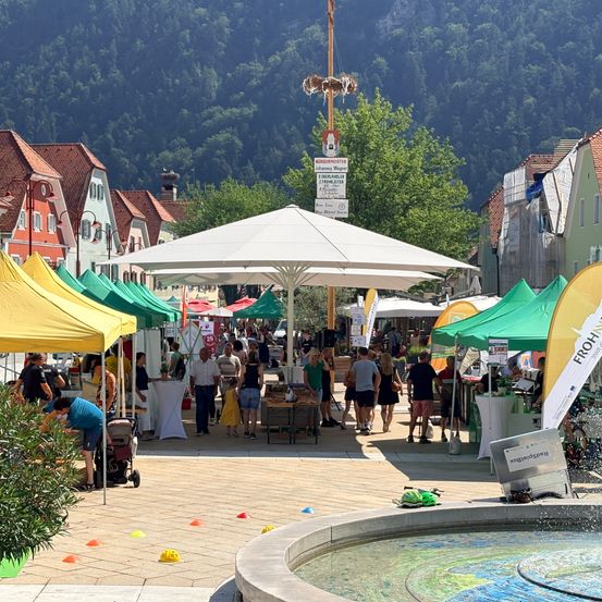 Ein geschäftiger Outdoor-Markt mit bunten Zelten, einkaufenden Menschen und einem Brunnen im Vordergrund. Berge und Gebäude sind im Hintergrund sichtbar.