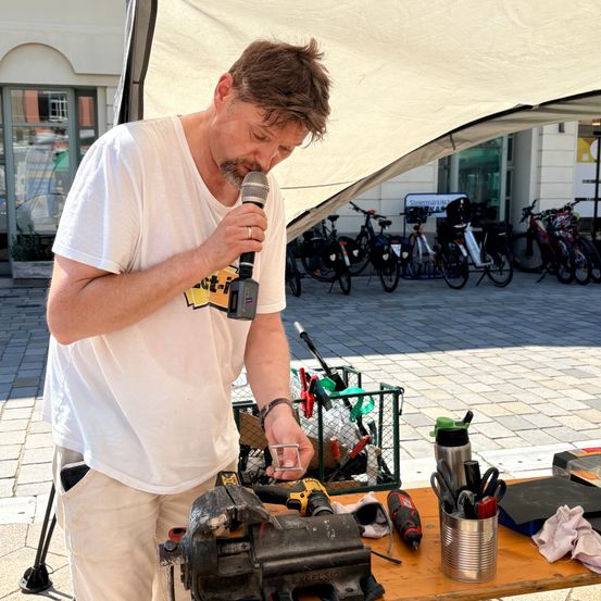 Ein Mann in einem weißen T-Shirt und khakifarbenen Hosen spricht in ein Mikrofon und hält ein Werkzeug in der Hand. Hinter ihm steht ein Tisch mit Werkzeugen und einem Fahrrad.