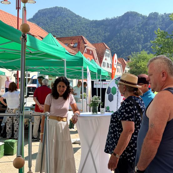 Eine Frau steht an einem Tisch mit einer Topfpflanze, lächelt und unterhält sich mit zwei Männern in einer Marktumgebung mit grünen Zelten und Bergen im Hintergrund.