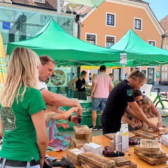 Eine Gruppe von Menschen arbeitet an einer Freiluftveranstaltung an Holzbrettern. Sie benutzen Werkzeuge und scheinen etwas zu bauen. Ein grünes Zelt ist im Hintergrund zu sehen.