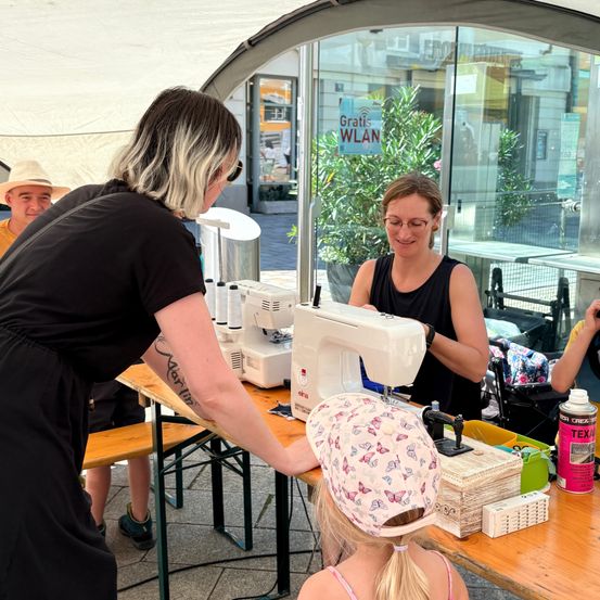 Eine Frau lehrt ein junges Mädchen, wie man eine Nähmaschine bei einer Veranstaltung im Freien benutzt.