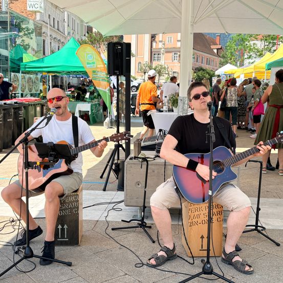 Zwei Männer spielen Gitarren auf einer Bühne auf einem Freimarkt, mit einer Menschenmenge, die vorbeigeht, und Ständen im Hintergrund.