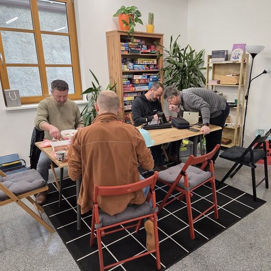 Vier Männer sitzen um einen Tisch in einem Raum mit Bücherregalen und Pflanzen, einer schaut auf einen Laptop, während ein anderer einen Becher hält.