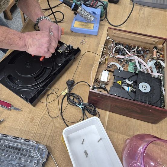 Eine Person arbeitet an einem demontierten elektronischen Gerät auf einem Holztisch, mit verschiedenen Werkzeugen und Komponenten um sie herum.
