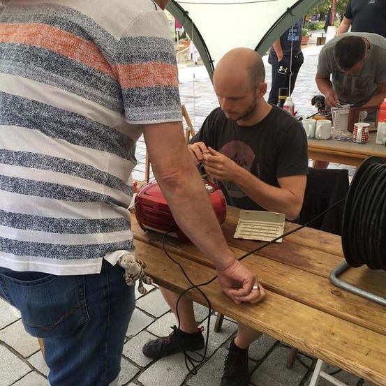 Ein Mann in einem schwarzen T-Shirt arbeitet an einem Projekt an einem Tisch in einer Außenumgebung. Ein anderer Mann in einem gestreiften Hemd steht neben ihm.