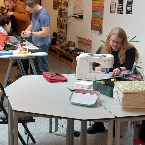 Eine Gruppe von Menschen arbeitet in einem Raum an verschiedenen Aufgaben. Eine Frau benutzt eine Nähmaschine, während ein Mann an einem Tisch mit verschiedenen Werkzeugen arbeitet. Ein anderer Mann steht und schaut sich etwas an.