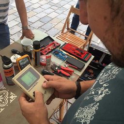 Ein Mann hält einen Nintendo Game Boy mit grünem Bildschirm, umgeben von verschiedenen Werkzeugen und Geräten auf einem Tisch.