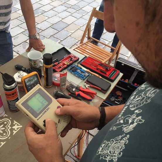 Ein Mann hält einen Nintendo Game Boy mit grünem Bildschirm, umgeben von verschiedenen Werkzeugen und Geräten auf einem Tisch.