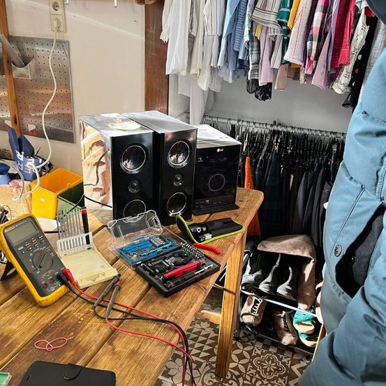 Ein überfüllter Raum mit einem Holztisch, auf dem elektronische Geräte, Lautsprecher und Werkzeuge stehen. Eine Person in einer blauen Jacke steht in der Nähe.