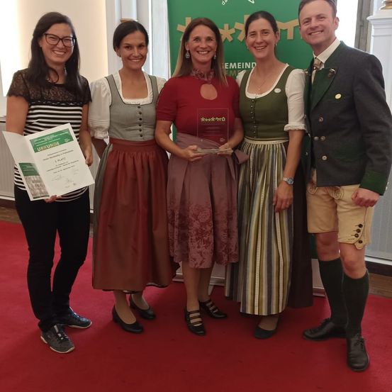 Fünf Personen stehen auf einem roten Teppich und lächeln für ein Foto. Eine Frau hält ein Zertifikat und eine andere ein Pokal. Sie sind in traditioneller Kleidung gekleidet. Hinter ihnen ist ein grüner Banner.