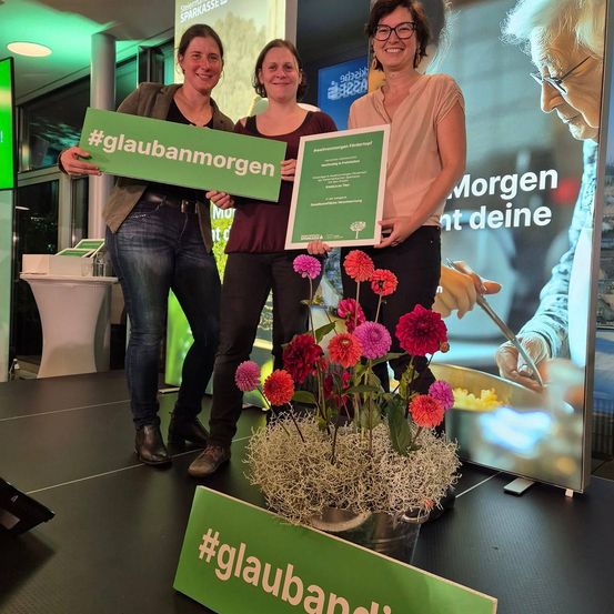 Drei Frauen stehen auf einer Bühne und halten ein grünes Schild mit dem Hashtag #glaubabanmorgen und einem Zertifikat. Im Vordergrund steht eine große Blumenarrangement. Dahinter zeigt eine große Leinwand eine ältere Person.