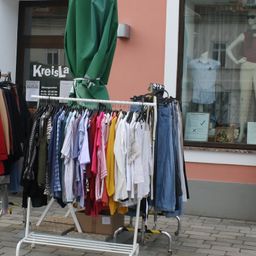 Ein Kleiderständer mit einer Vielzahl von Kleidungsstücken, die vor einem Geschäft mit dem Schriftzug 'KreisLa' ausgestellt sind. Im Schaufenster sind Mannequins mit Sonnenbrillen und lässiger Kleidung zu sehen.