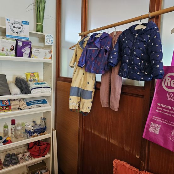 Ein Zimmer mit einem weißen Regal voller Babyprodukte und Kleidung, die an einer Holzwand hängt.