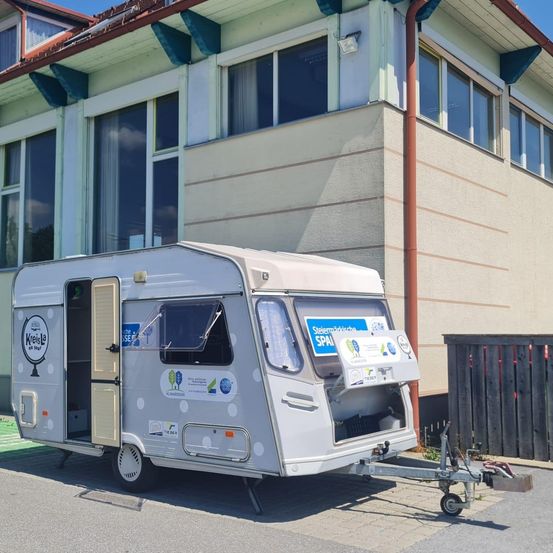 Ein weißer Wohnwagen mit Punktmuster steht vor einem Gebäude mit Glasfenstern und einem roten Dachrinnen.