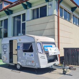 Ein weißer Wohnwagen mit Punktmuster steht vor einem Gebäude mit Glasfenstern und einem roten Dachrinnen.