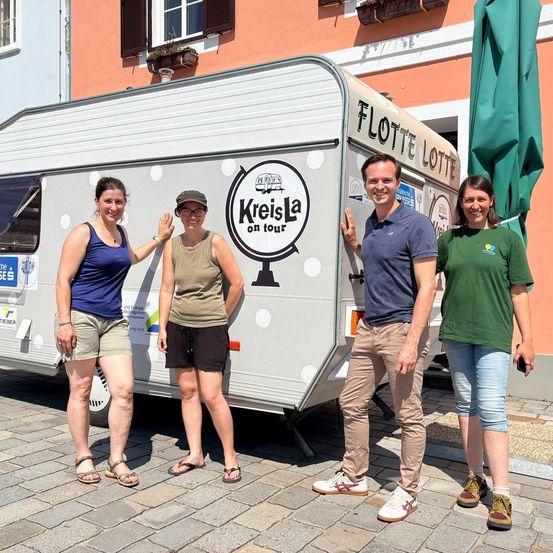 Vier Personen stehen vor einem weißen Anhänger mit den Worten 'Kreisla on tour' und einem Globus-Logo. Sie lächeln und posieren für ein Foto.