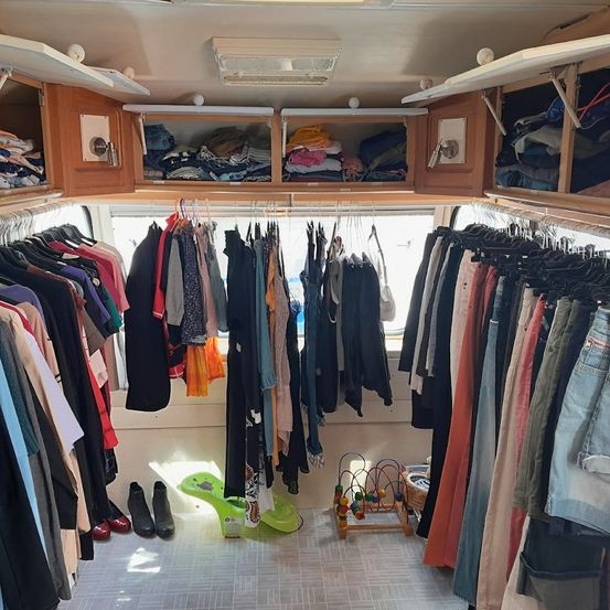 Ein gut organisierter Kleiderschrank, gefüllt mit verschiedenen Kleidungsstücken, ordentlich an Kleiderbügeln und Regalen aufgehängt. Der Raum hat Holzschränke, ein Fenster und einen gefliesten Boden.