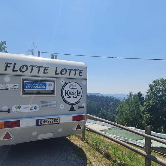 Die Rückseite eines Wohnmobils namens Flotte Lotte, geparkt an einer Straße mit einer malerischen Aussicht auf Berge und Bäume.
