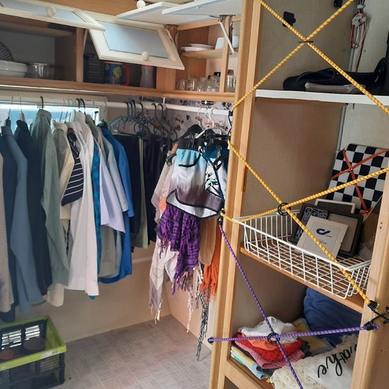 Ein kleiner, organisierter Kleiderschrank mit Kleidung an einer Seite und Regalen mit verschiedenen Gegenständen auf der anderen.