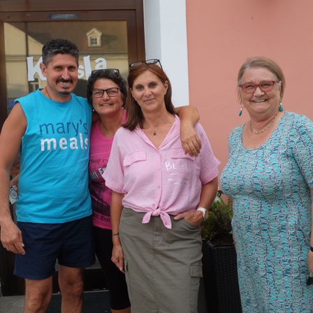 Vier lächelnde Erwachsene stehen vor einem Gebäude mit Glasfenstern. Der Mann trägt ein blaues T-Shirt mit den Worten 'mary's meals'.