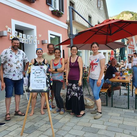 Fünf Personen stehen vor einem Café und lächeln für ein Foto. Die Gruppe besteht aus drei Frauen und zwei Männern. Die Frau auf der linken Seite hält ein Schild mit einer Papierkette, während die anderen beiden Frauen Halsketten tragen. Ein roter Schirm befindet sich hinter ihnen.