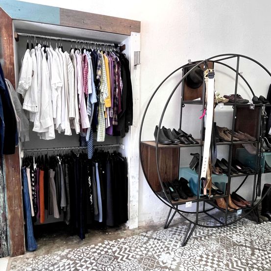 Innenansicht eines Boutique-Ladens mit einer Reihe von Hemden an einem Kleiderständer und einem runden Ständer mit Schuhen, einer Mütze und Taschen.