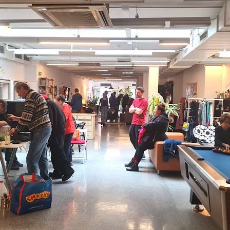 Ein Innenbereich mit mehreren Personen, einem Mann in einem roten Shirt, der mit verschränkten Armen steht, einer Frau, die auf einer Couch sitzt, einem Tisch mit einer Tasche, einem Billardtisch und Kleidung, die an einem Kleiderständer hängt.