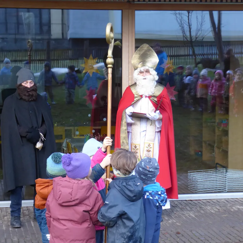 Ein Mann, der als Sankt Nikolaus verkleidet ist, steht neben einer Person in einem schwarzen Umhang und hält einen Stab. Mehrere Kinder sind um sie herum versammelt. Die Szene spielt sich vor einem Gebäude mit Glaswänden ab, die eine Reflexion von Menschen und einem Baum zeigen.