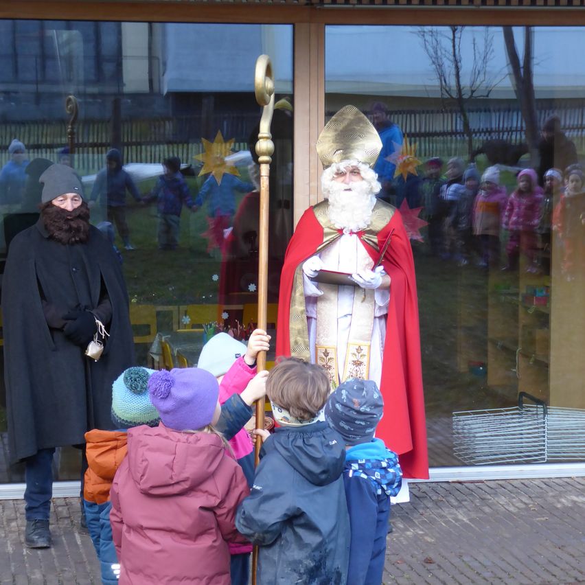 Ein Mann, der als Sankt Nikolaus verkleidet ist, steht neben einer Person in einem schwarzen Umhang und hält einen Stab. Mehrere Kinder sind um sie herum versammelt. Die Szene spielt sich vor einem Gebäude mit Glaswänden ab, die eine Reflexion von Menschen und einem Baum zeigen.