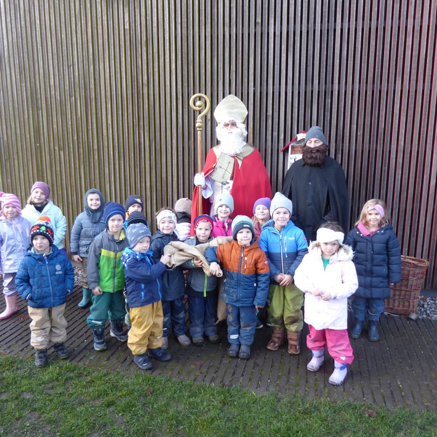 Eine Gruppe von Kindern steht vor einer Holzwand und posiert mit einer Person in Santa-Claus-Verkleidung und einer anderen in einem schwarzen Umhang.