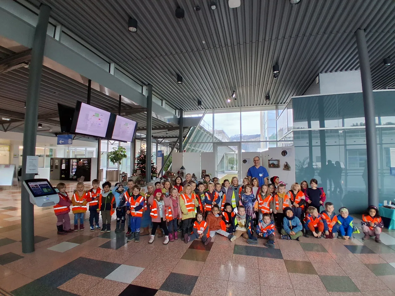 Eine Gruppe von Kindern in Warnwesten posiert für ein Foto in einer Lobby, mit Monitoren und einem Weihnachtsbaum im Hintergrund.