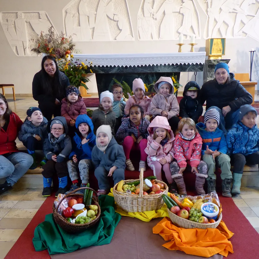 Eine Gruppe von Kindern und Erwachsenen posiert für ein Foto in einer Kirche mit Körben voller Obst und Gemüse auf dem Boden vor ihnen.
