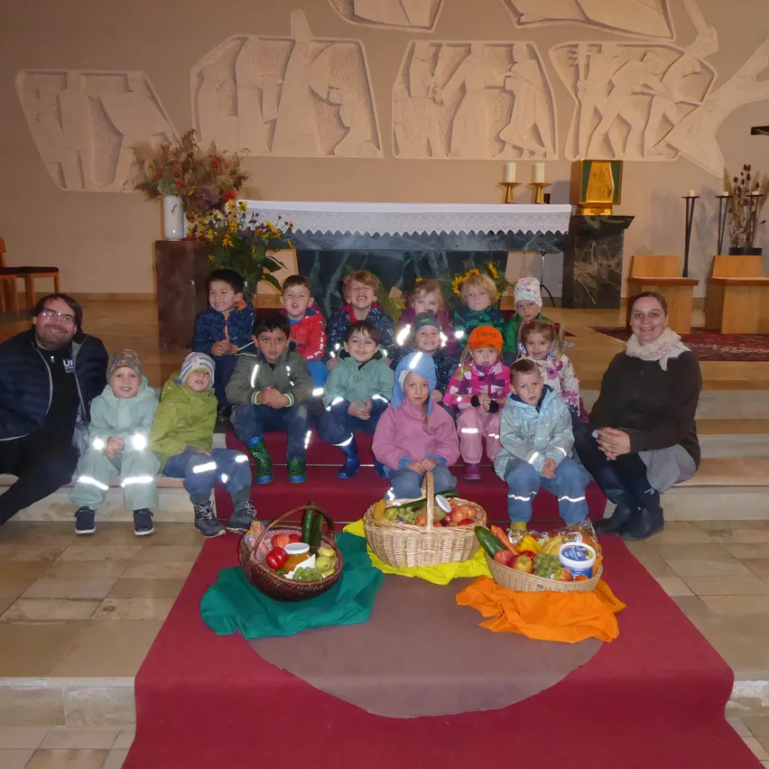 Gruppenfoto von Kindern und Erwachsenen, die in einer Kirche posieren. Zwei Erwachsene sitzen auf den Stufen, und die Kinder sind vor einem Korb mit Obst und Gemüse aufgereiht. Dahinter steht ein Kirchenaltar mit Kerzen und einem Kreuz.