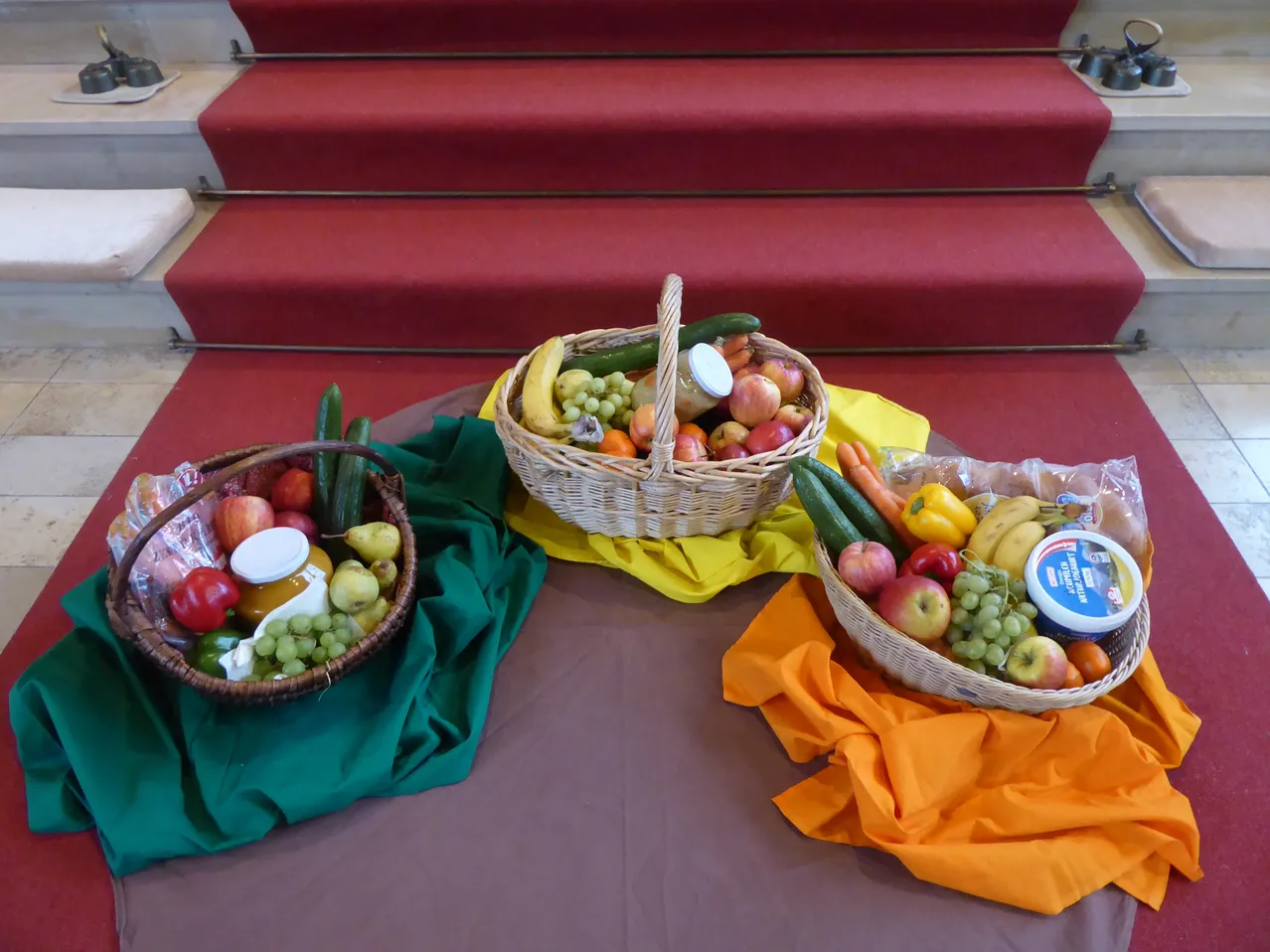 Drei Weidenkörbe mit verschiedenen Obst- und Gemüsesorten auf einem Tisch, einer mit einem gelben Tuch, einer mit einem grünen Tuch und einer mit einem orangefarbenen Tuch, vor dem Hintergrund einer rot Teppich bezogenen Treppe.