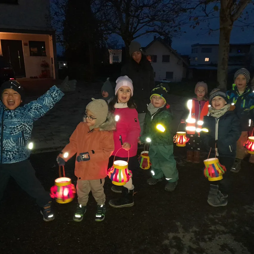 Eine Gruppe von Kindern mit leuchtenden Laternen posiert nachts für ein Foto. Eine Frau steht hinter ihnen in der Nähe eines Hauses.