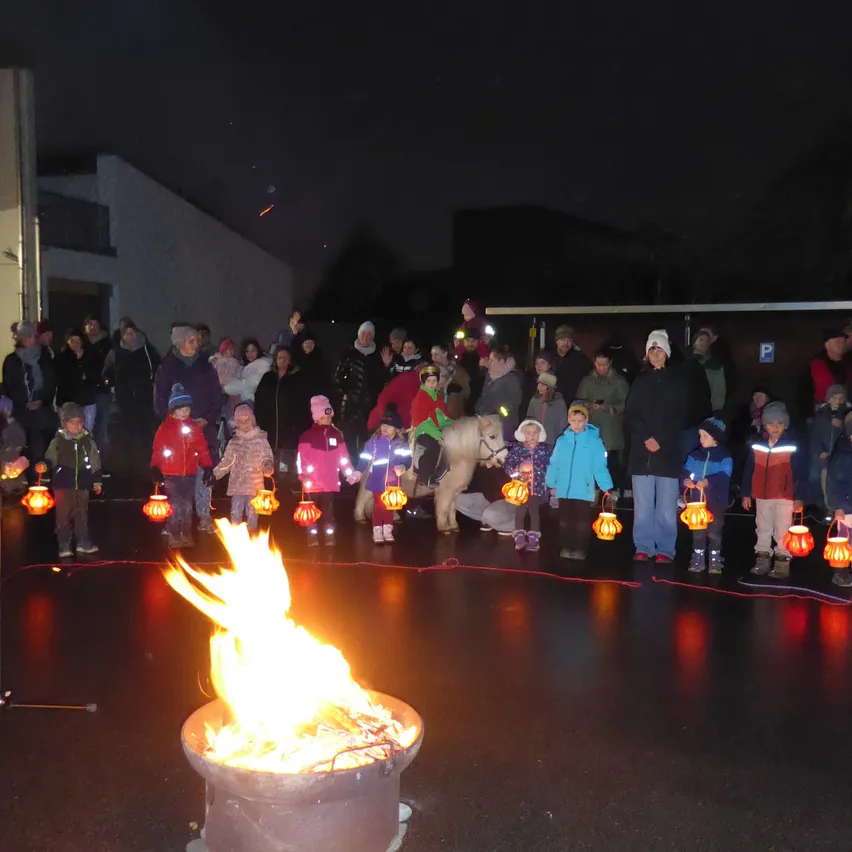 Eine Menschenmenge, darunter Kinder, versammelt sich um ein Lagerfeuer und hält Laternen in einer nächtlichen Umgebung.