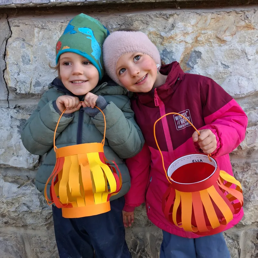 Zwei Kinder stehen nebeneinander und halten Papierlaternen in den Händen. Sie lächeln und scheinen für ein Foto zu posieren. Hinter ihnen ist eine Steinmauer.